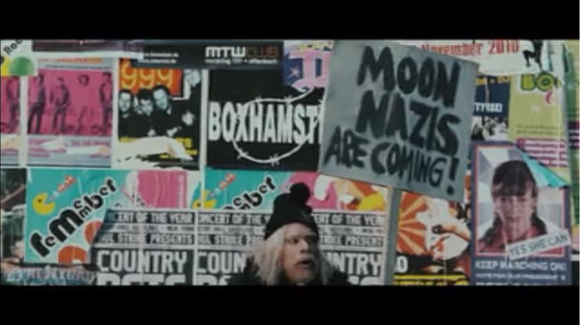 A man with shaggy hair and beard stands in front of a wall, holding up a sign that reads "Moon Nazis are coming!"
