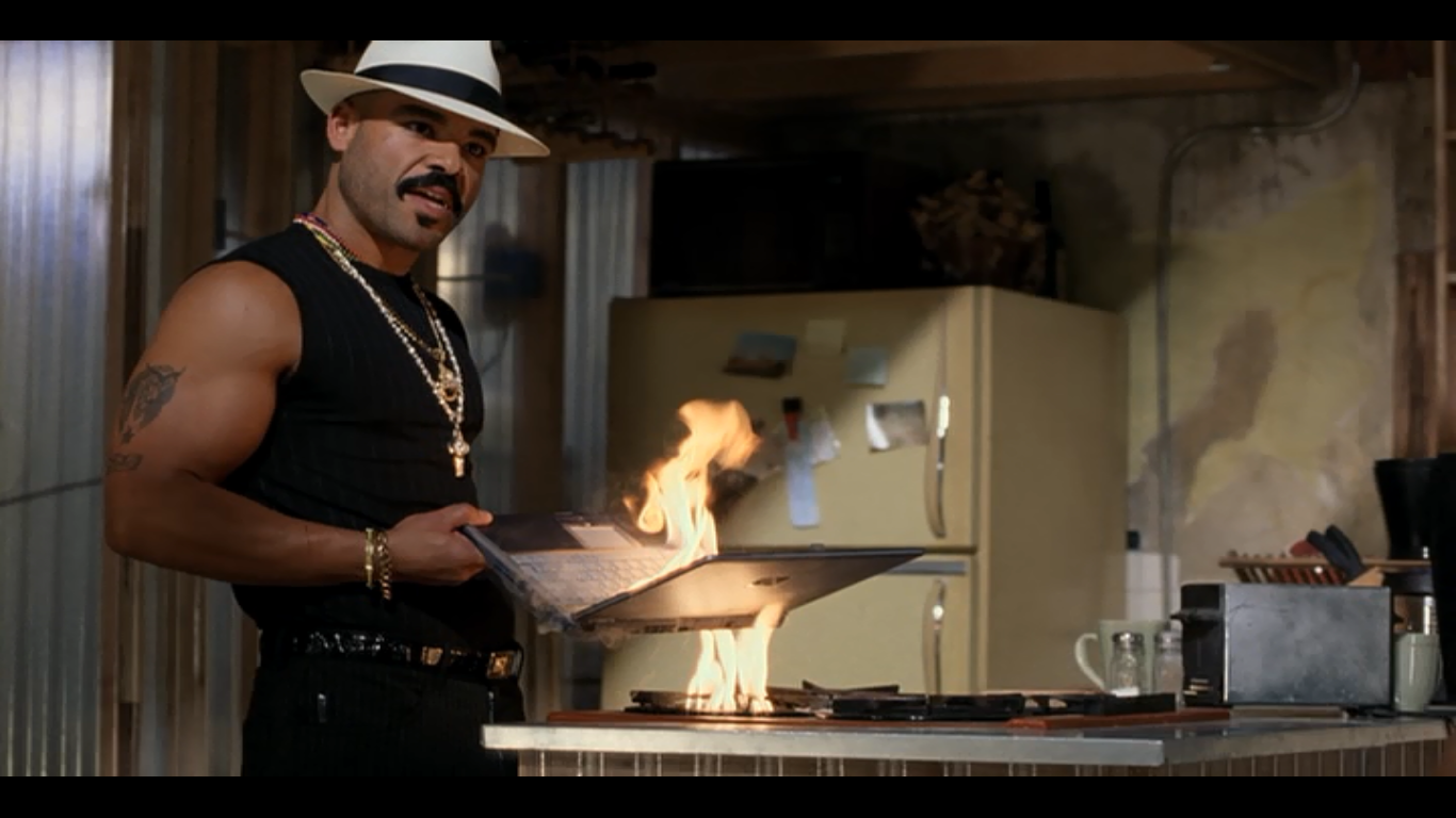a man with a fedora and several gold chains holds a laptop over an open flame in a kitchen