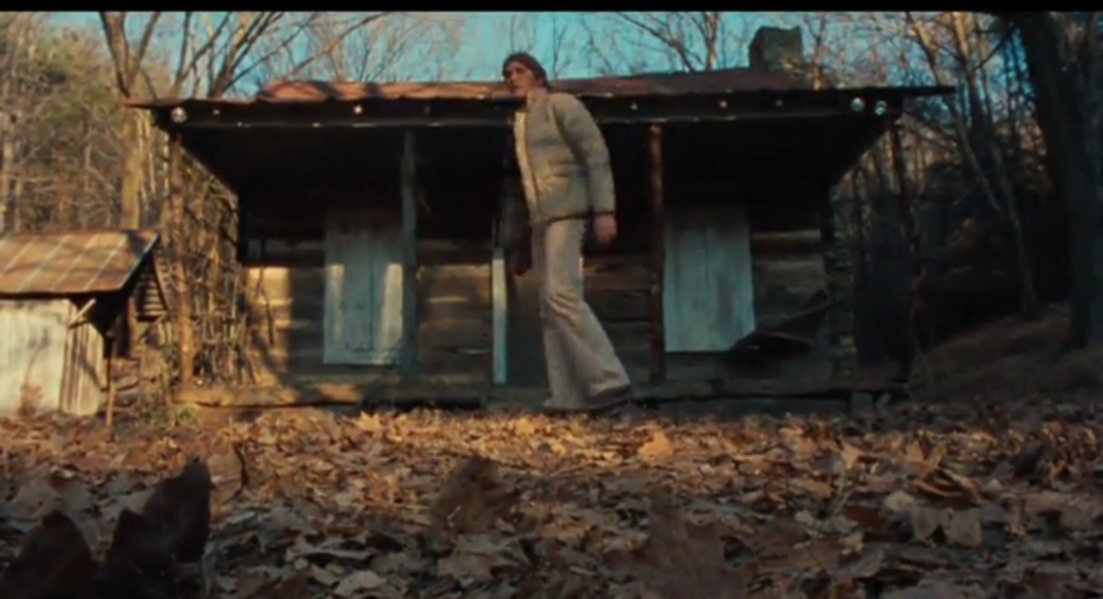 a young man stands outside of a small cabin with boarded-up windows in the woods