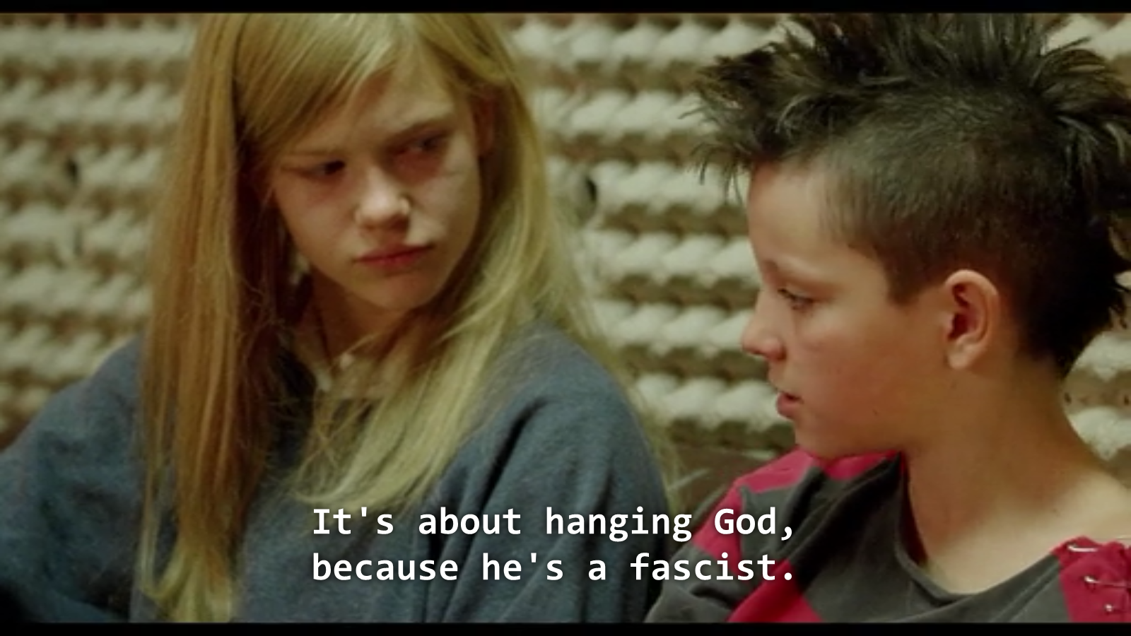 a teenage girl with long blonde hair looks skeptically at a girl with a mohawk who tells her "It's about hanging God, because he's a fascist"