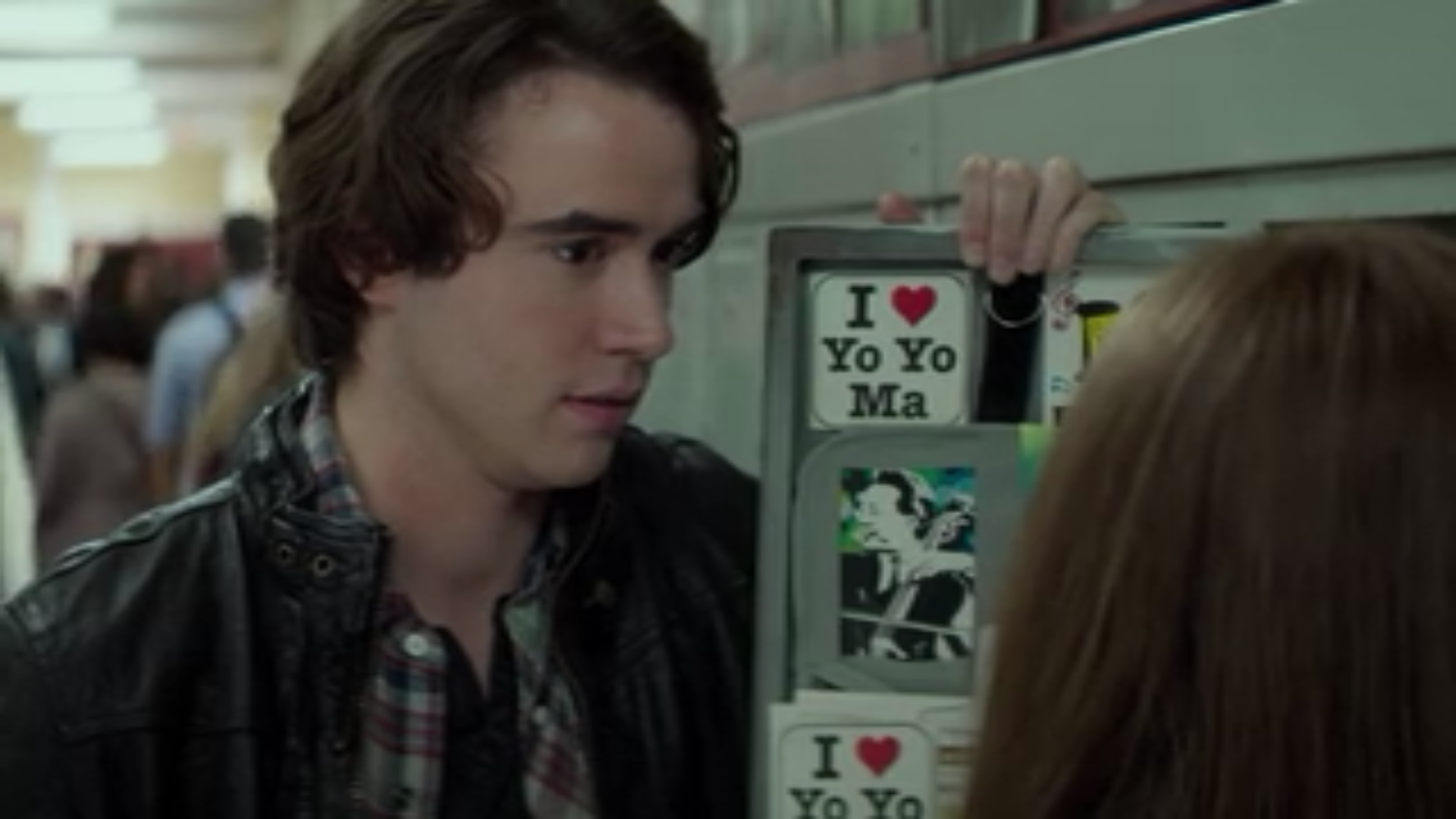 A teen boy stands by a girl's open locker, which is decorated with multiple stickers that read "I Heart Yo Yo Ma."