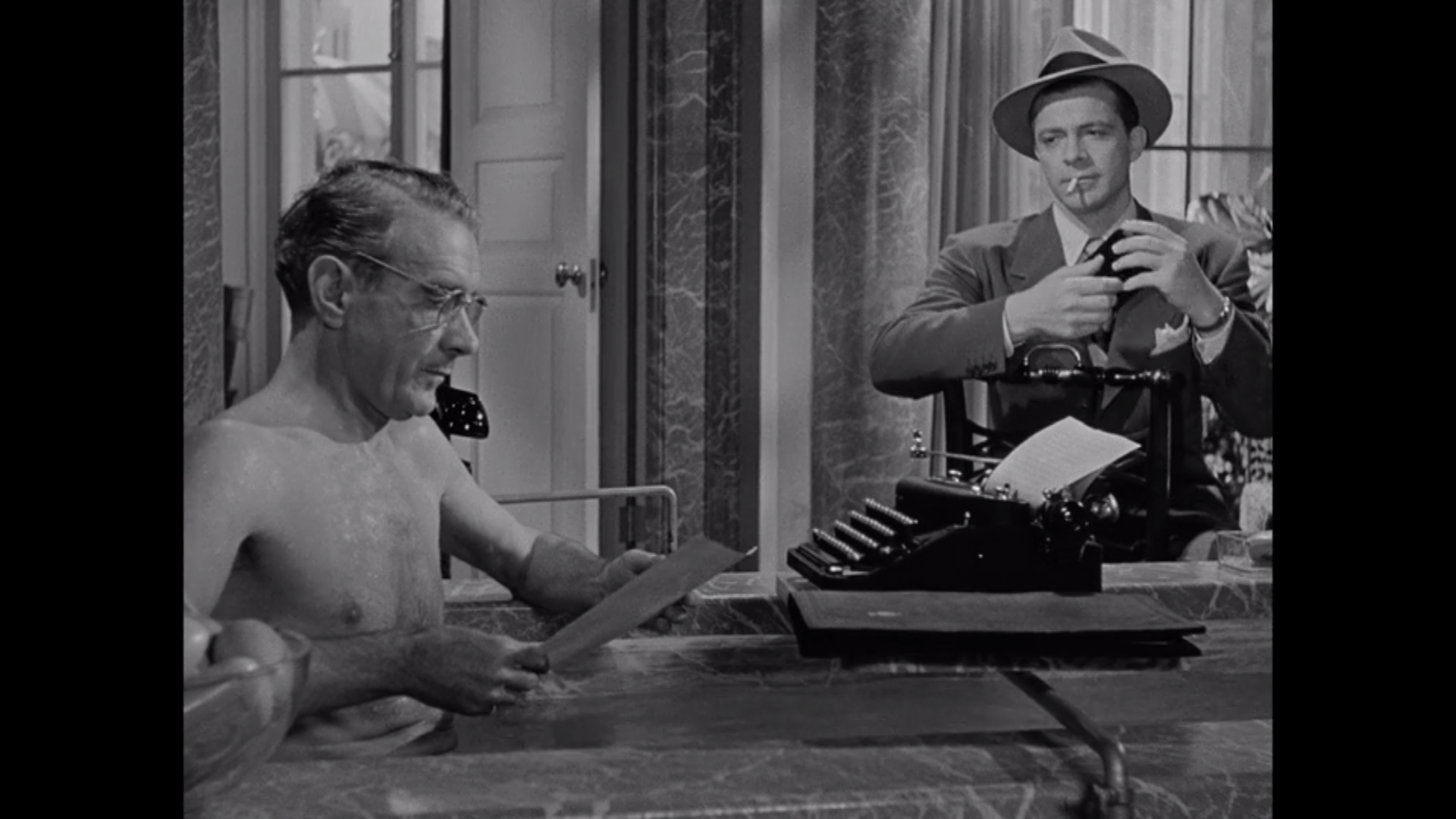 A man in a fedora smokes a cigarette, facing a man in a bathtub who is using a typewriter.