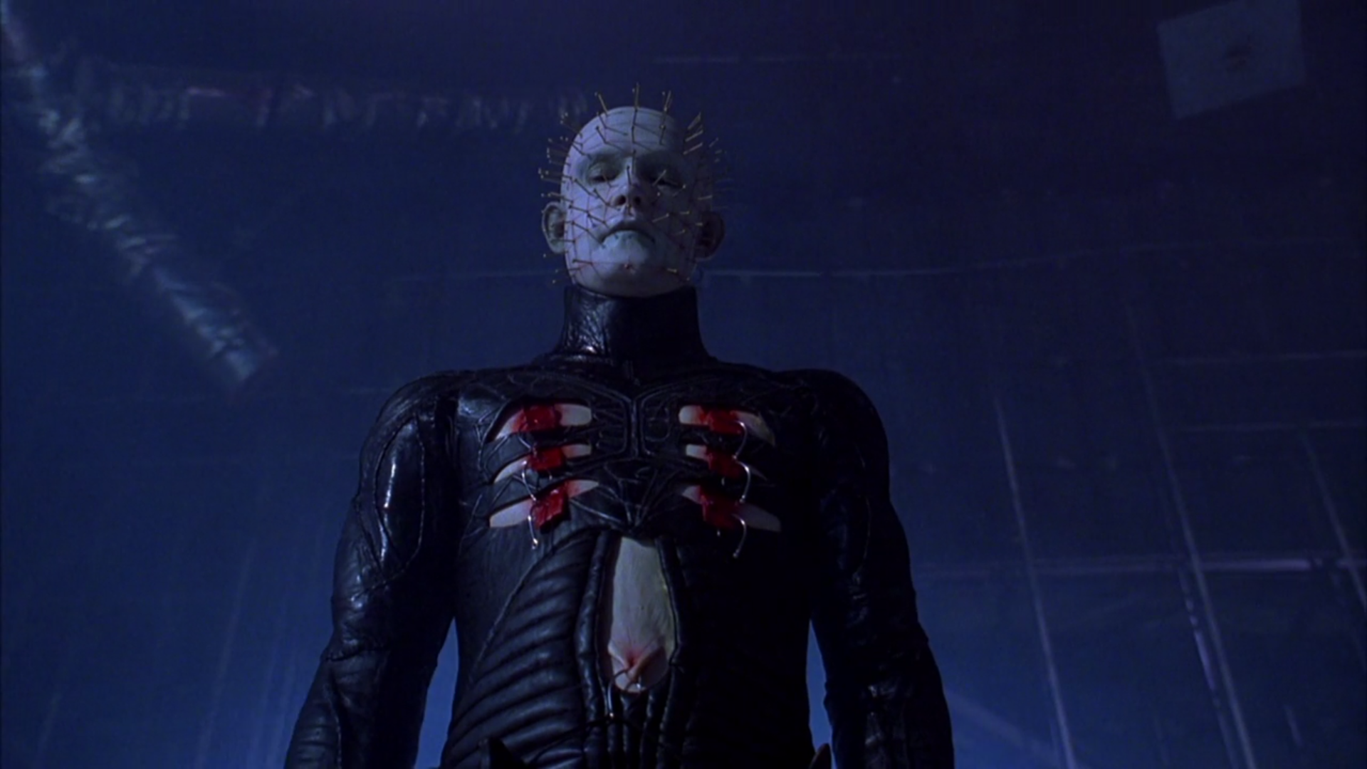 Pinhead, a demon wearing all-leather and with pins sticking out of his face and skull, looms above the camera, looking down.