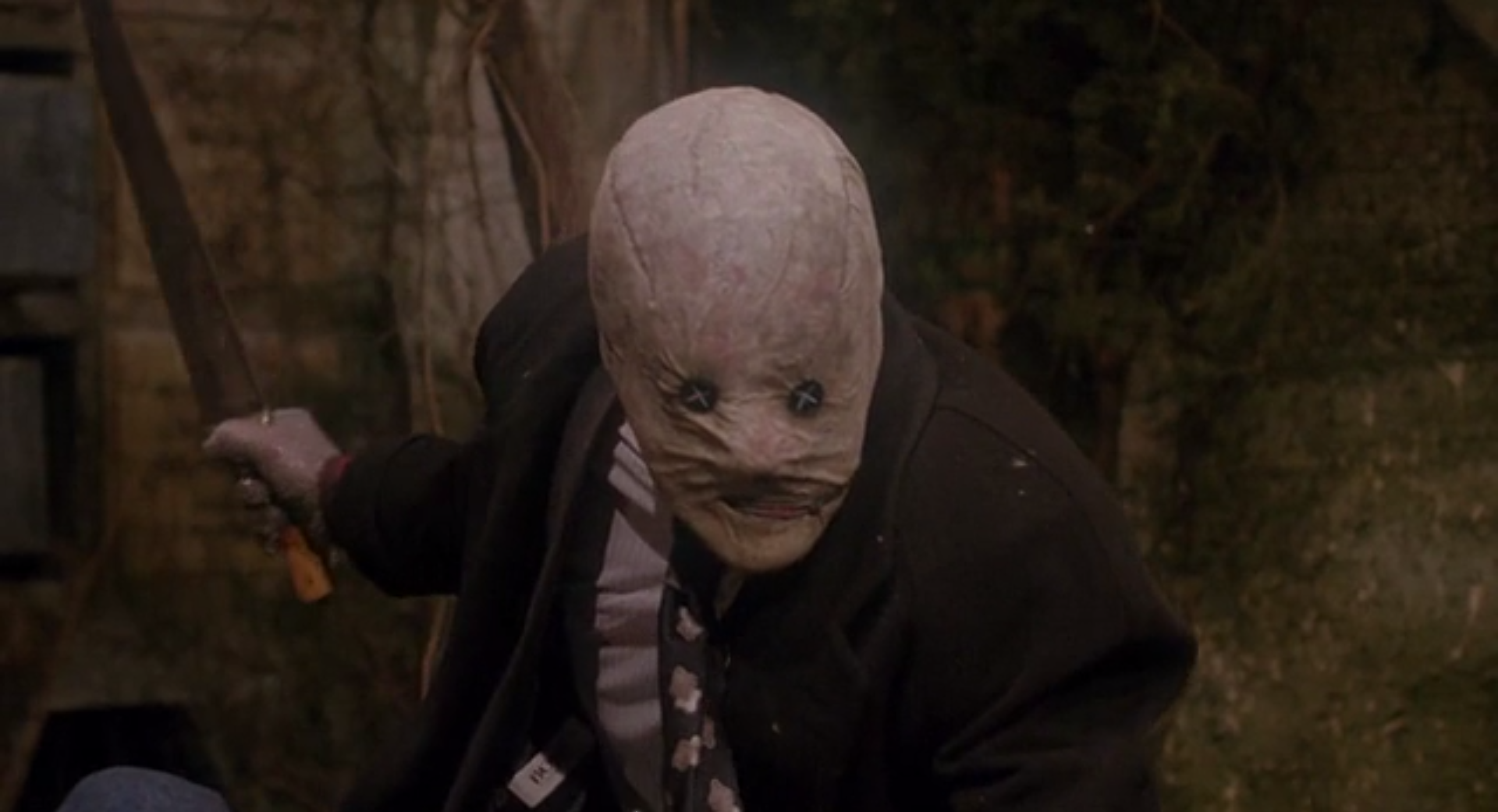 A man swings a large knife, wearing a mask that fits his face closely and has buttons sewn over the eyes.
