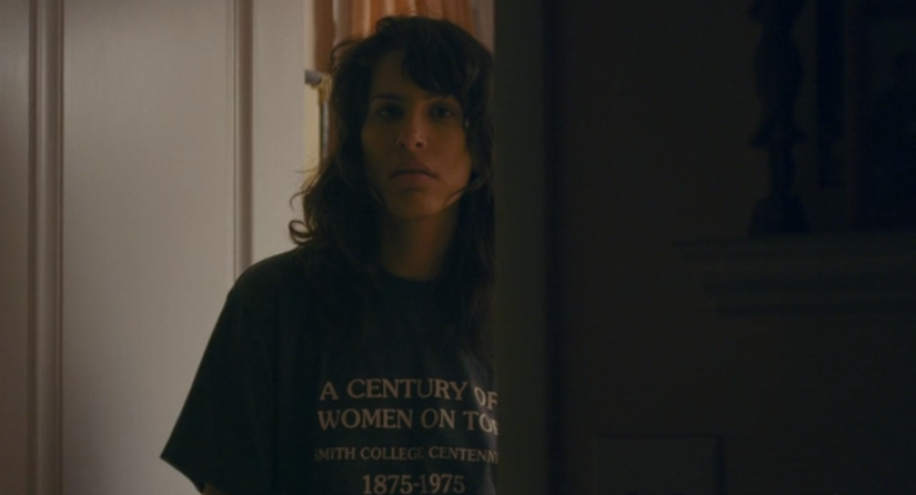 a woman stands in a dimly lit doorway wearing a t-shirt that reads "A century of women on top: Smith College centennial: 1875-1975"