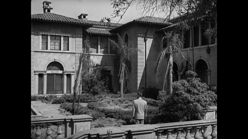 a man stands in front of a large Spanish-style mansion