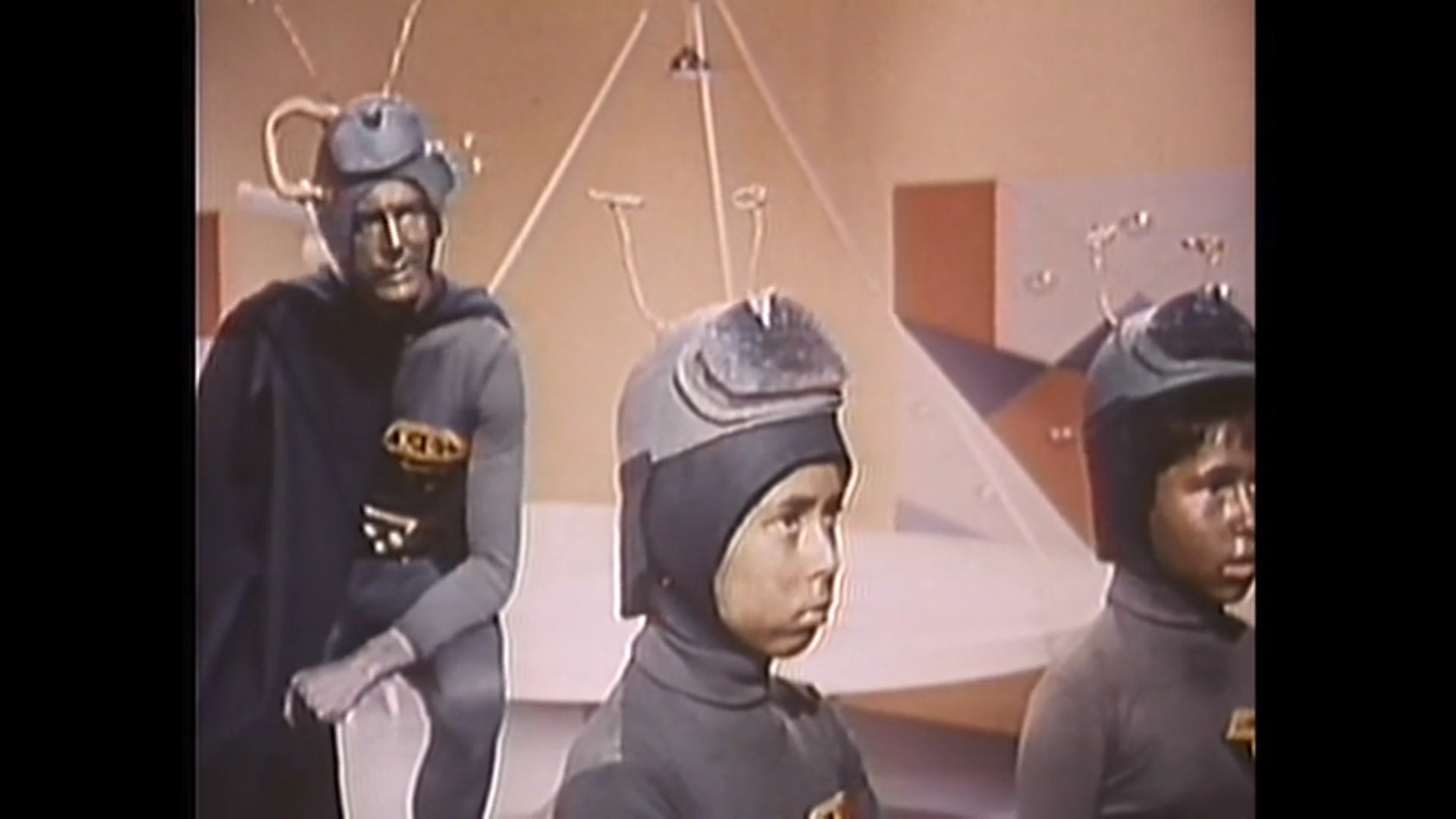 two boys in matching blue costumes and helmets stand while a man dressed identically sits in the background
