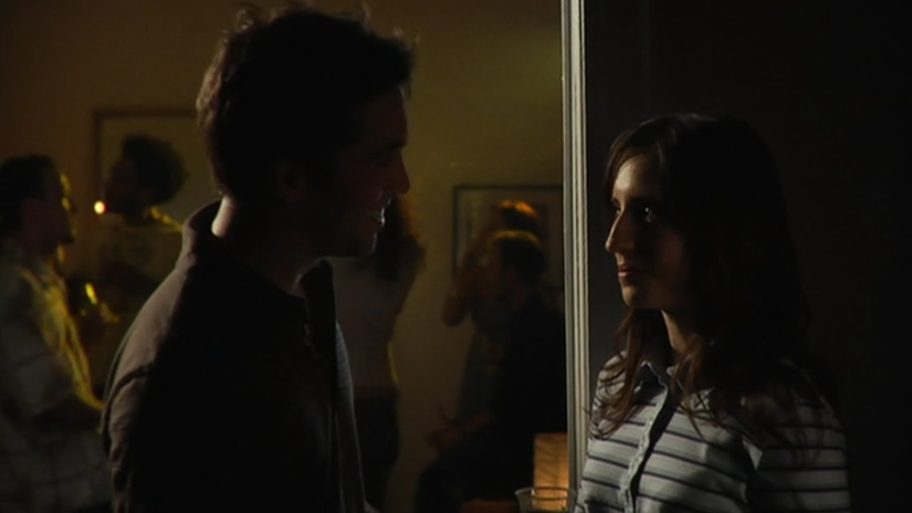 a woman wearing a polo shirt talks to a man at a party in an apartment