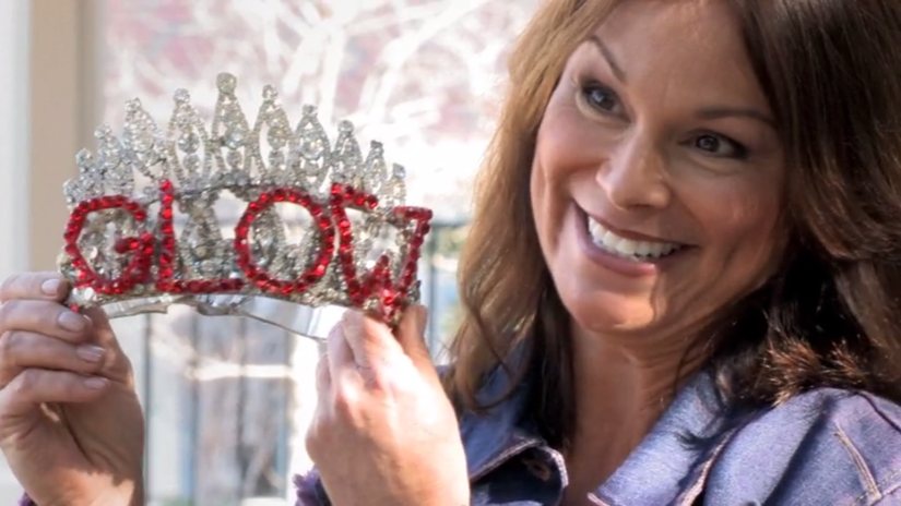 a woman smiles, holding up a crown with the word "GLOW" written in red rhinestones
