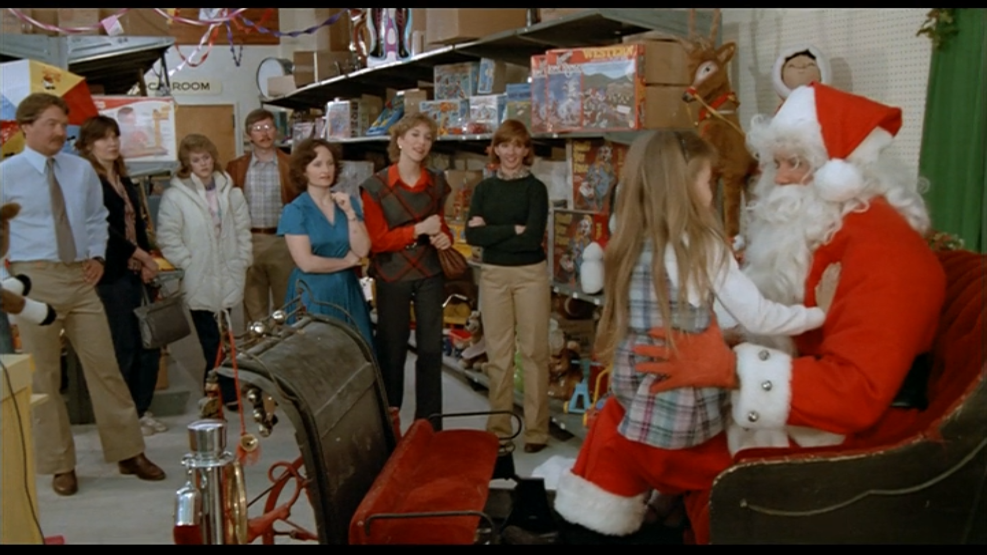a girl sits on the lap of a toy store Santa while a group of adults look on