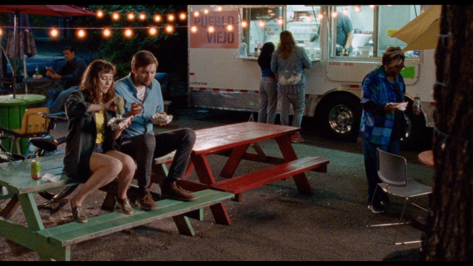 A man and woman sit next to each other on top of a picnic table, eating tacos from a taco truck parked in the background.