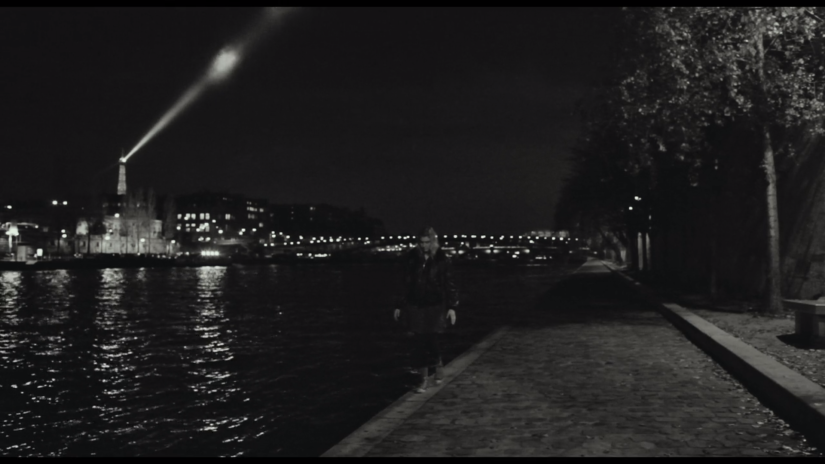A woman walks along the edge of the Seine River at night, the Eiffel Tower behind her in the distance