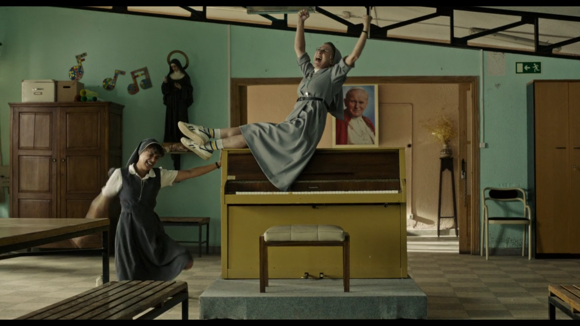 A nun sits on top of a piano, raising her hands skyward while a novice stands next to her, dancing.