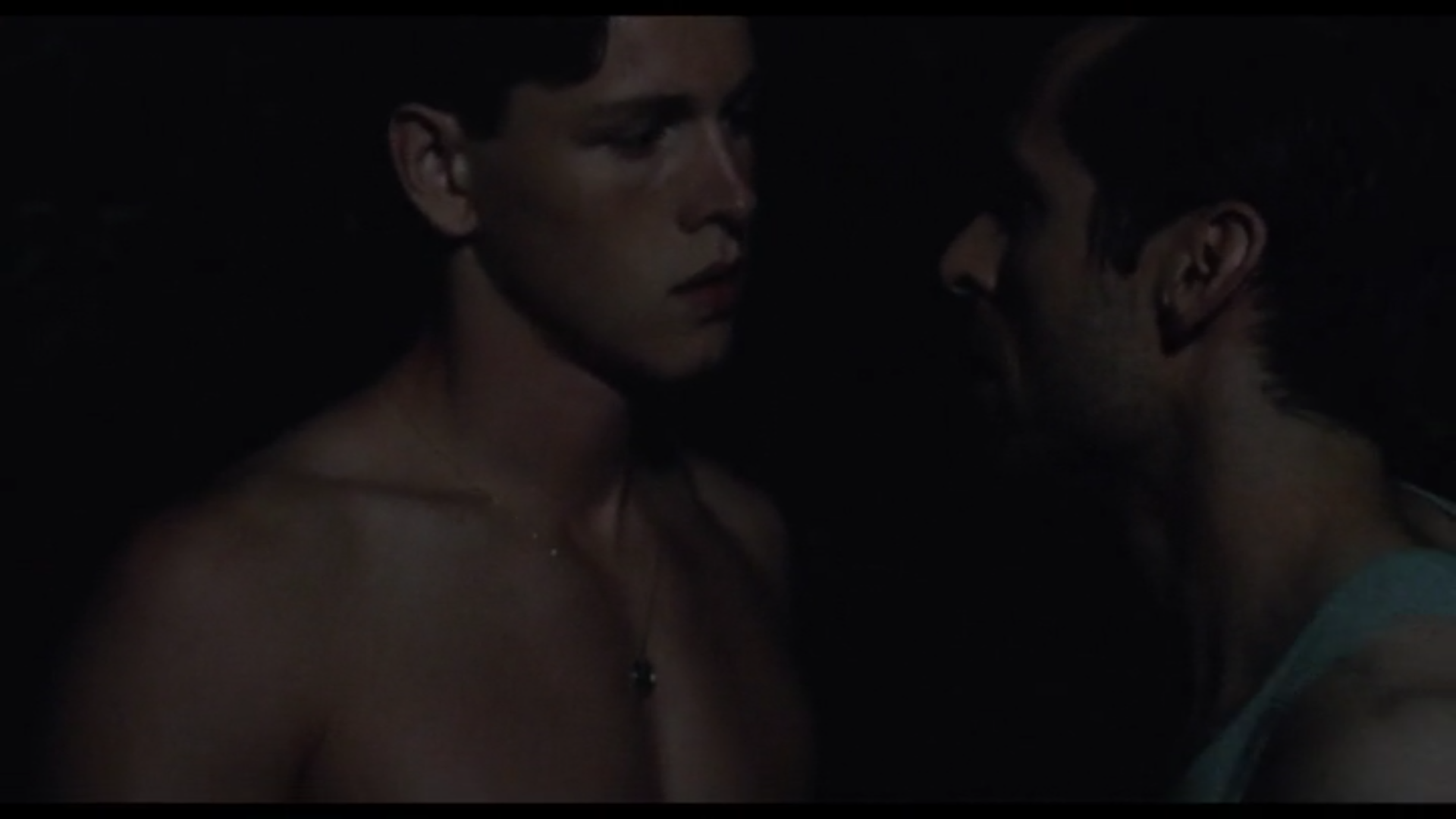 a shirtless man faces another man in the dark