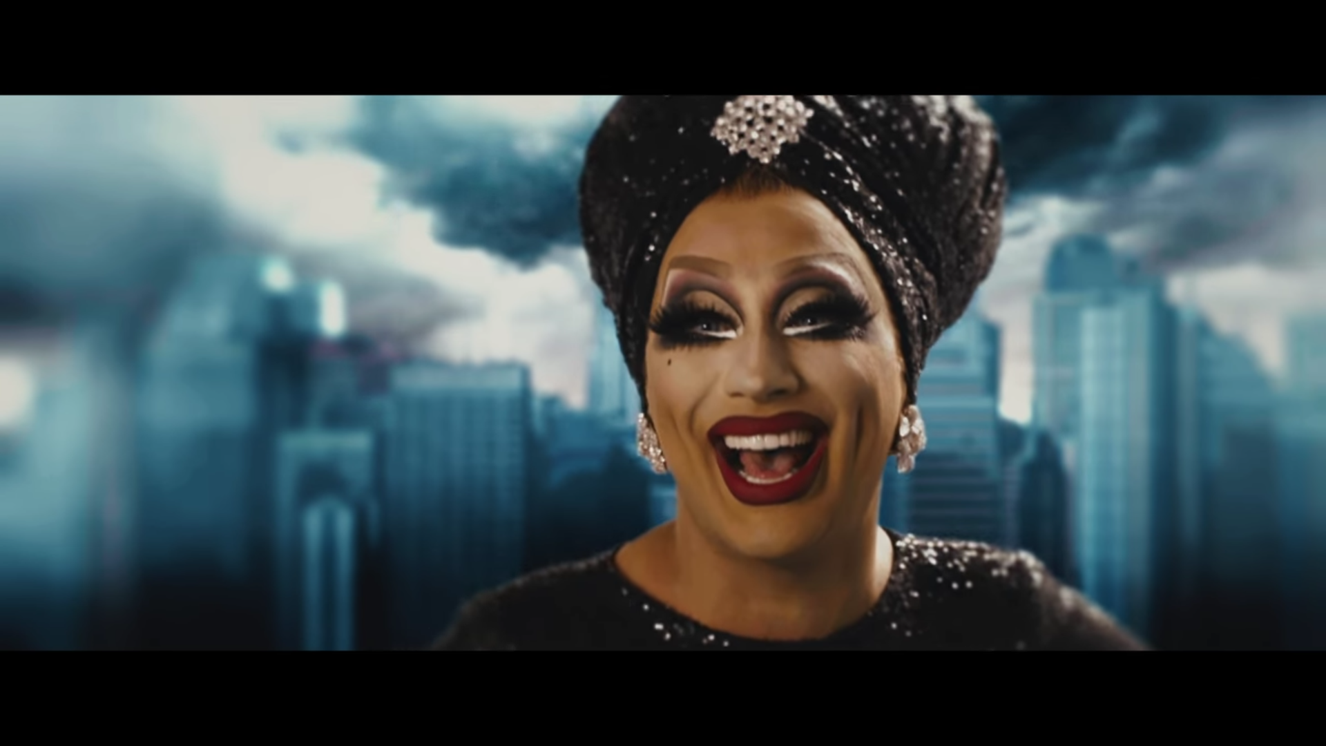 A drag queen wearing a sparkly turban laughs, a cloudy cityscape behind her.