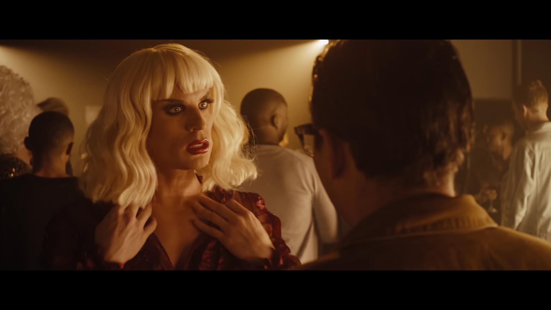 A man talks to a blonde drag queen at a bar.