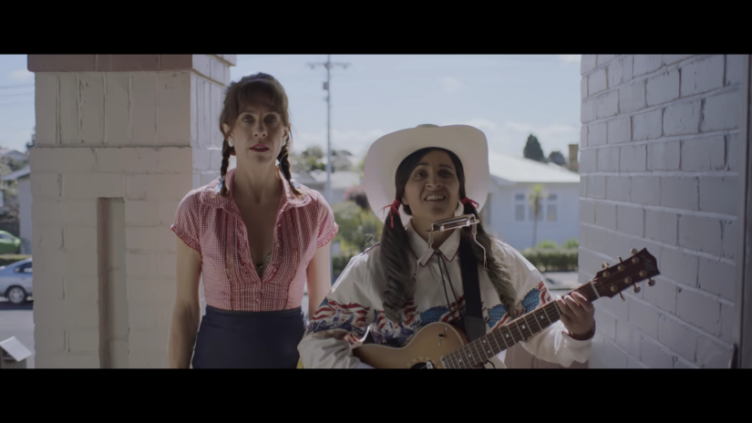 a woman with pigtail braids stands next to a woman in a cowboy hat who is playing the guitar and harmonica