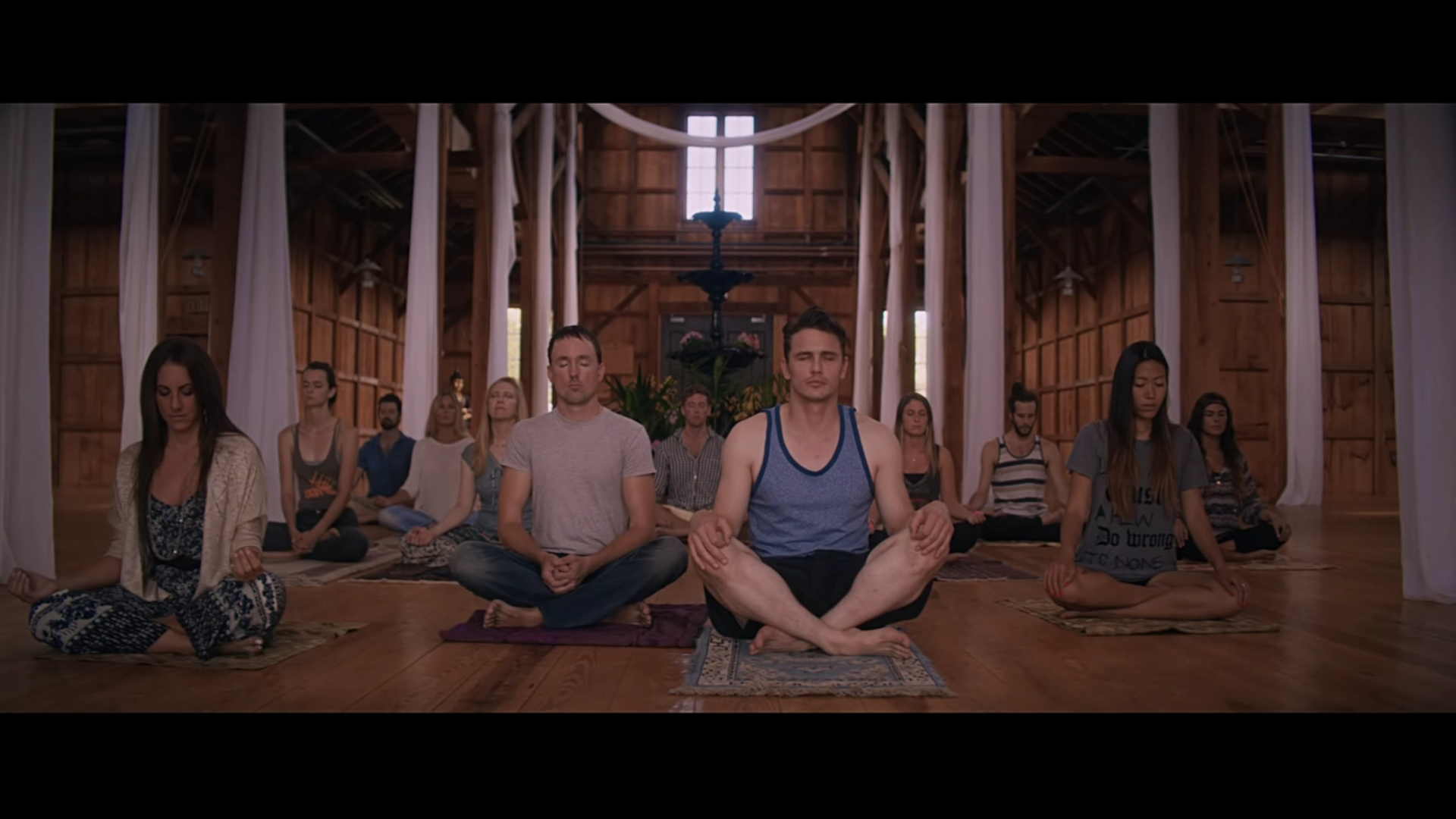 A group of people meditate on rugs in a large room with wooden floors and walls. They sit with crossed legs and eyes closed, and a man in a blue tank top and black shorts is the most prominent.