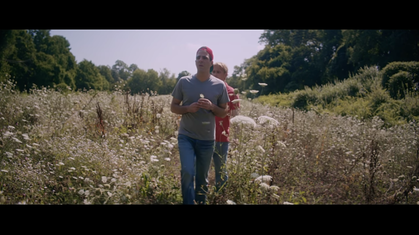 Two men walk through a meadow of white wildflowers. The man in front holds a flower and wears a backwards red baseball cap, blue t-shirt and jeans. The man behind him has blond hair, and wears a red t-shirt and jeans.