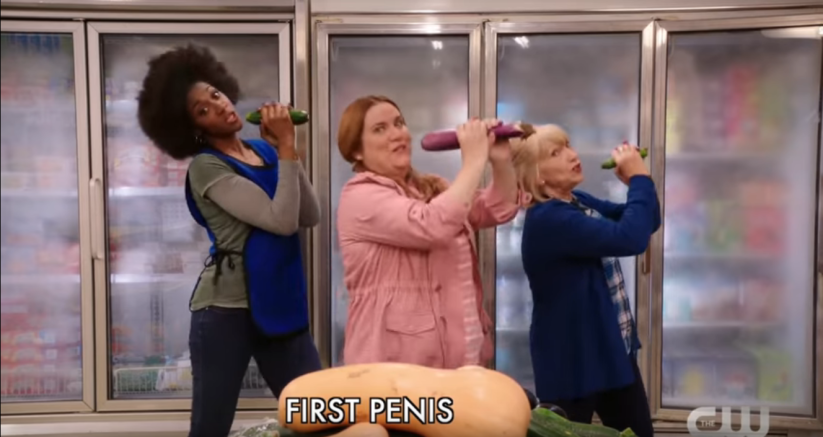three women holding vegetables as microphones stand in front of a large squash, singing the words "First penis"