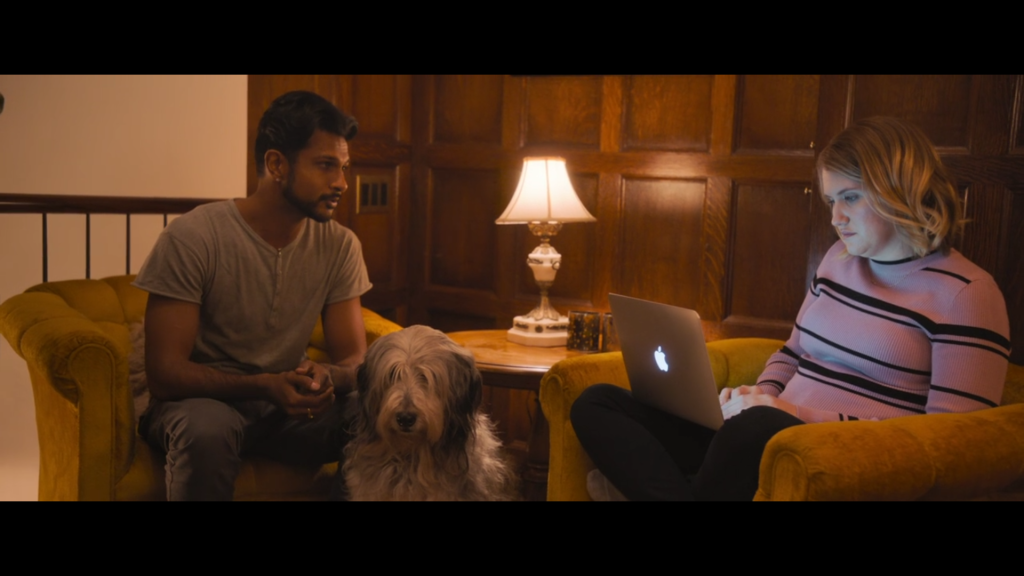 A woman sits in an armchair, working on a laptop. A man is looking at her, while a dog sits between them.
