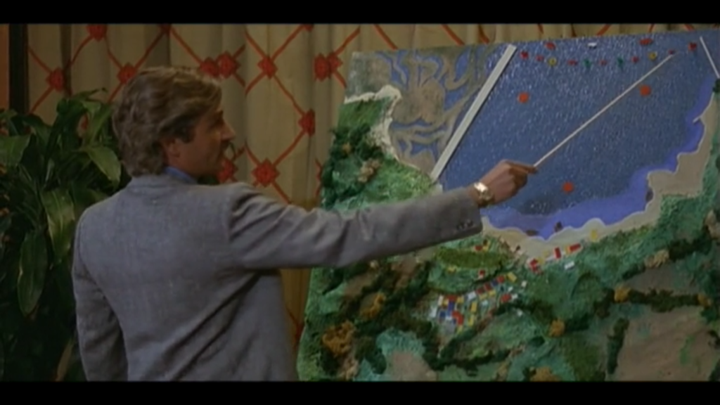A man in a suit with 1970s hair swept back uses a white pointer to gesture to boats on a map of a small bay.