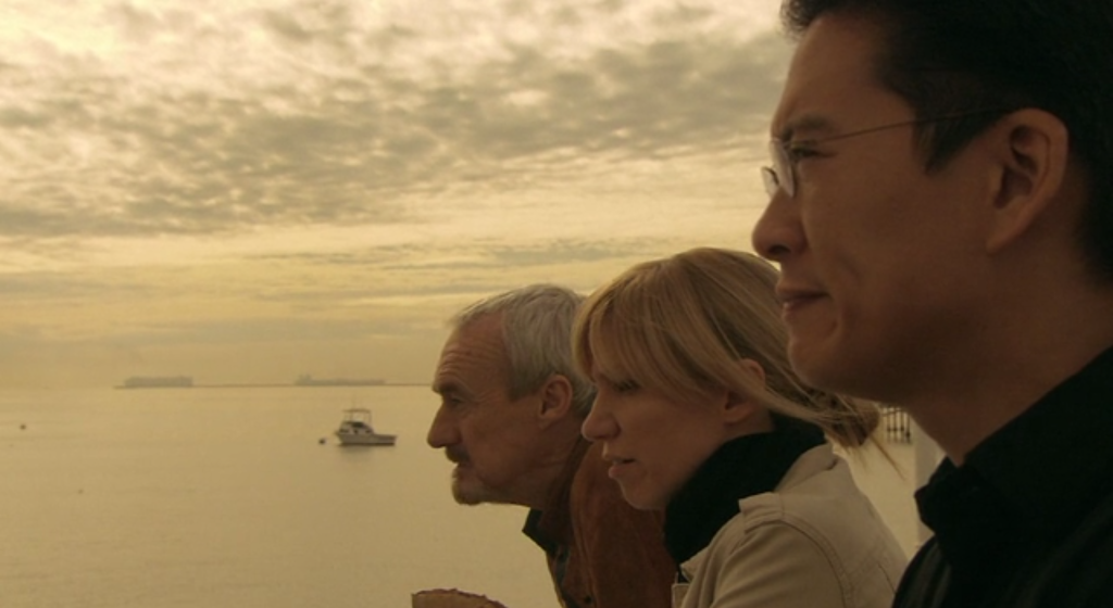 3 people look out towards the ocean: an elderly man with graying hair, a blonde woman with hair in a messy bun, and an Asian man wearing glasses.