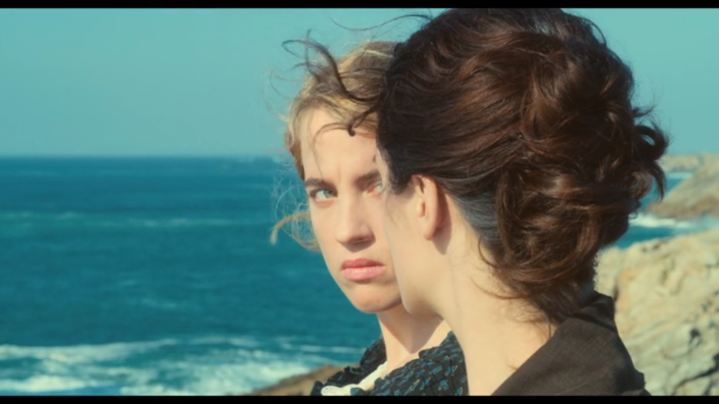 On a windswept beach, a blonde woman looks seriously at a dark-haired woman.