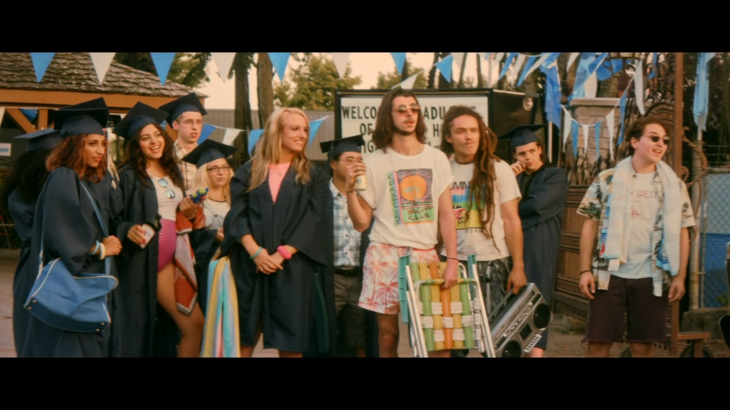 A group of teens in a mix of beachwear and graduation gowns stand at the entrance of a water park.