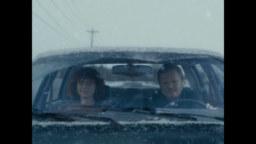 Jake, a man with blonde hair, drives along a snowy country road with Lucy as a passenger.