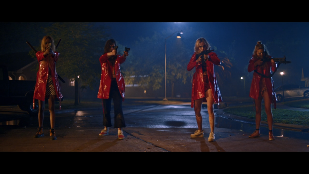 Dressed in red PVC trench coats, a group of 4 teen girls stand in a row, aiming firearms at an unseen group of boys.