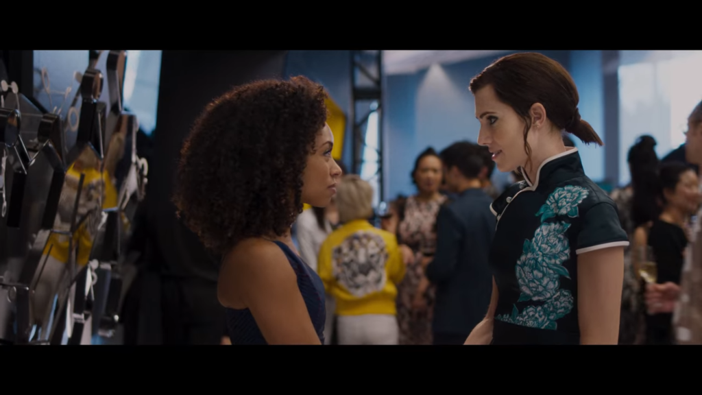 Lizzie, a young Black woman, converses with Charlotte, a young white woman. They are dressed formally, standing apart from groups of other people gathered for a reception.