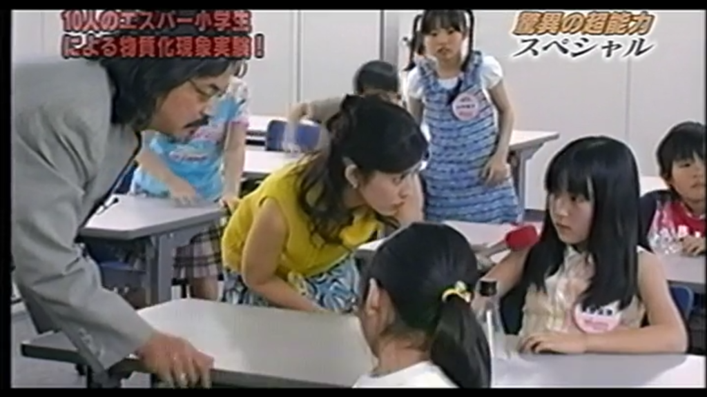 Two of the makers of a television show stand over the desk of a young student, Kana. She is seated, looking hesitant as a microphone is held to her face.