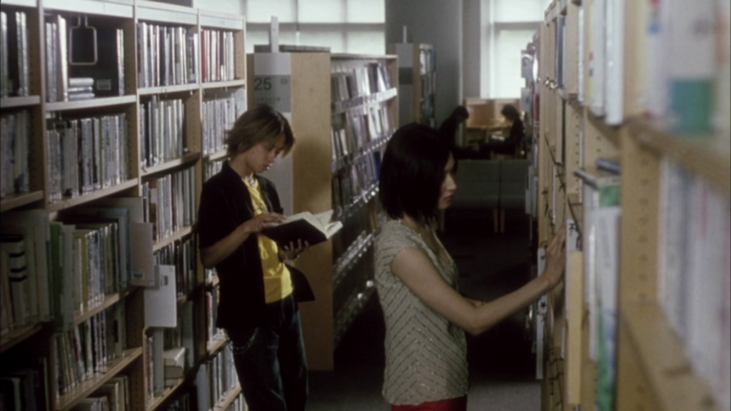 Harue, a young woman browsing the shelves in an academic library, stands facing away from Ryosuke, who is leaning against the shelves reading.