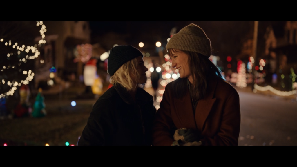 The character of Abby faces her girlfriend Harper as they hold hands at night, Christmas lights on the houses behind them. The two are arm-in-arm, smiling, and dressed warmly in coats and hats.