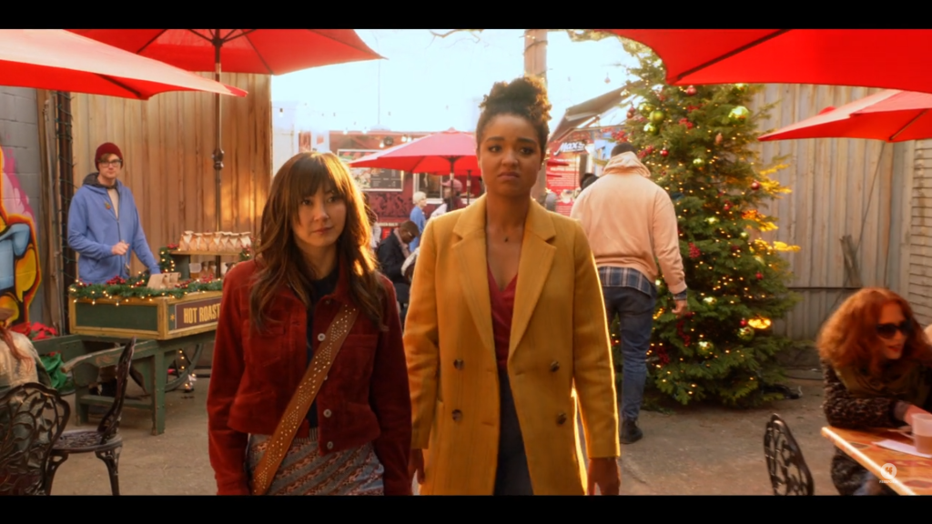 Kara, a young Asian-American woman with long dark hair walks next to Jess, a young African-American woman with her hair up. They are walking through an outdoor Christmas market with booths and tables set up under red umbrellas.