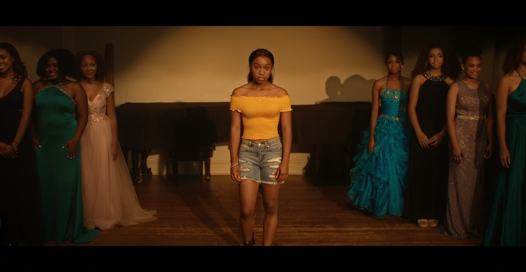 A teenage girl wearing a yellow shirt and ripped jean shorts stands onstage, teenage girls in formal gowns standing on either side of her.
