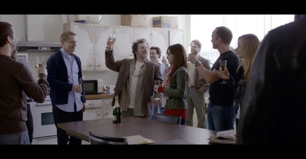 In a rundown staff lounge, a man leads a toast to another man, with around 10 other people joining in.