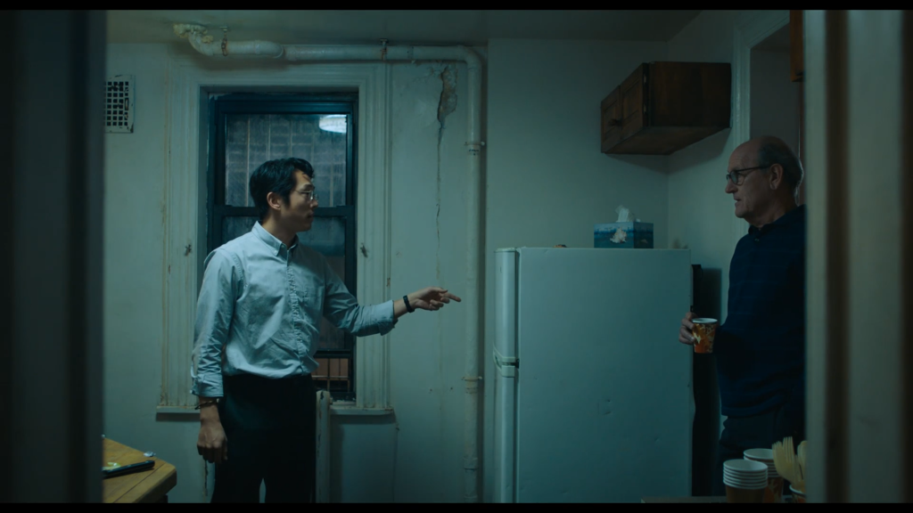 Two men stand in a dirty, aged kitchen, having a conversation.