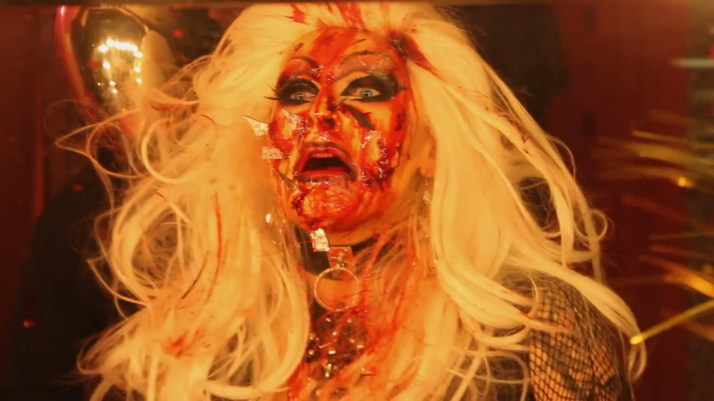 A drag queen wearing a long blonde wig gasps in horror at her blood face, pierced by shards of glass from a mirror.
