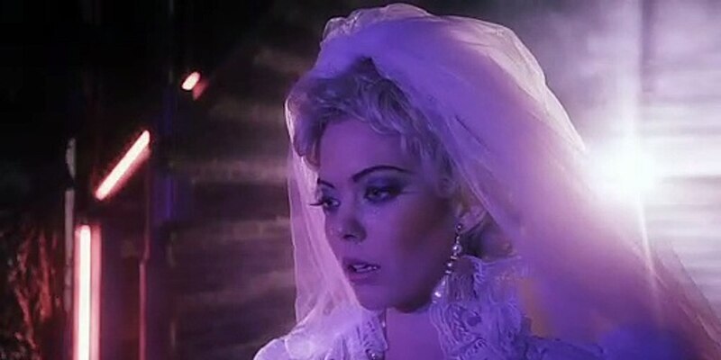 A blonde model wearing a wedding dress and veil walks in an alley, bright lights behind her backlighting the scene.