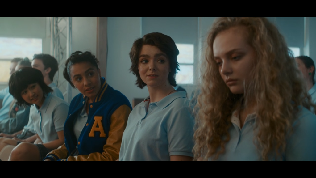 Three teenage girls look expectantly at another teen girl, whose eyes look glazed over as she stares down at the floor.